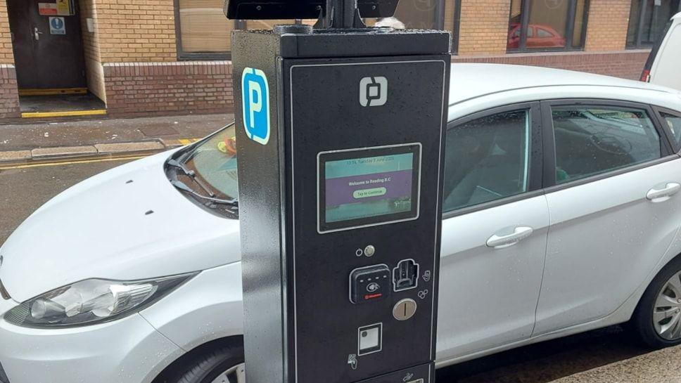 A black parking machine with a contactless panel on it. There's a white car parked on the side of the road behind it.