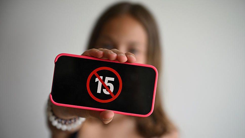 A teenager holds her smartphone displaying a logo "ban under 15". The phone screen is black with a crossed out 15 in the middle of it. The person is blurred in the background and the phone is held in front of them facing the camera.