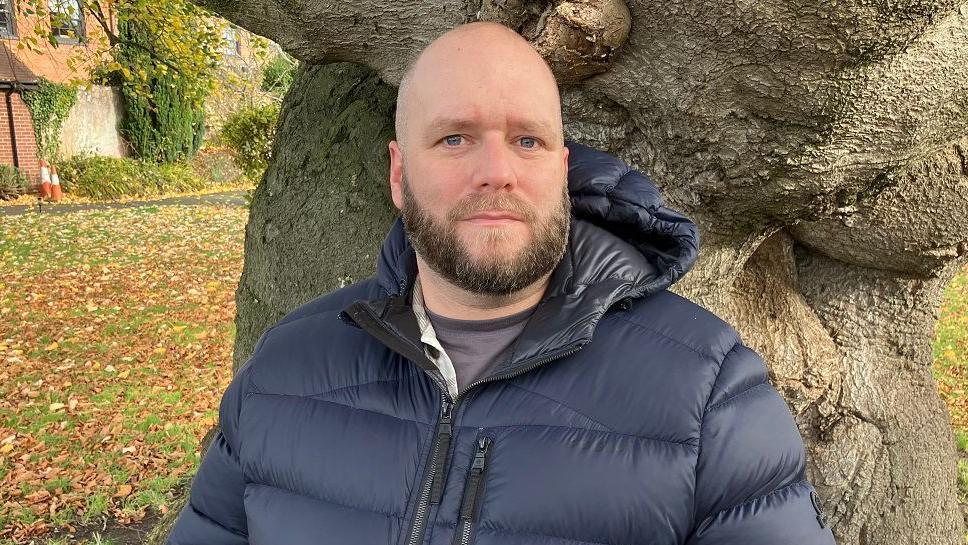 Pete White is an RAF veteran who left the armed forces in 2017. He is bald with a beard and is wearing a puffer jacket and standing in front of a tree, looking into the camera. There is a grassed area behind him.