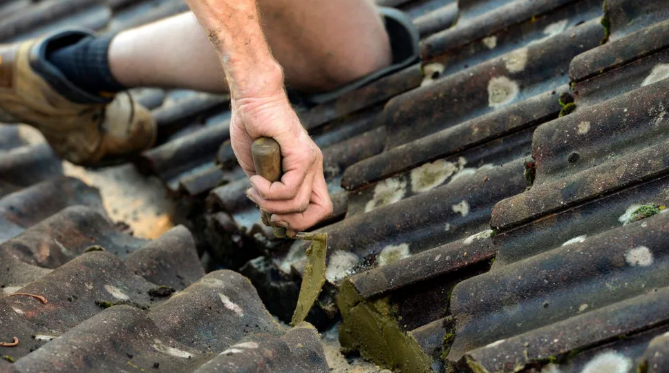 A corrugated tiled roof is being repaired by a man whose knee and arm can be seen. He is holding a trowel which is scraping one of the tiles.