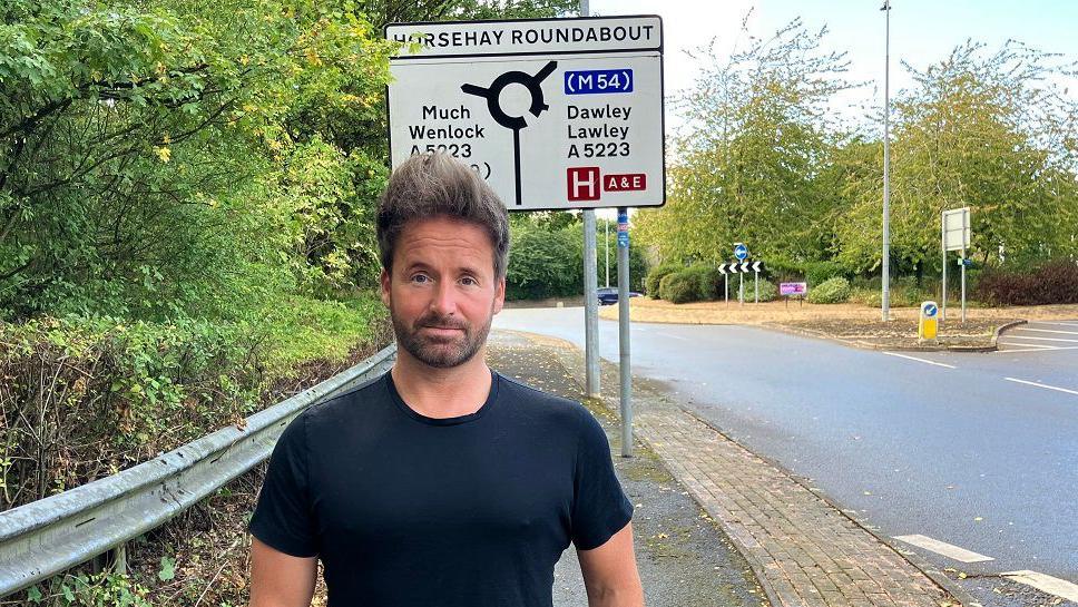 Councillor Ian Preece standing in front of a road sign in Horsehay in Telford where there has been incidents of car cruising on Sunday nights, which have been reported by residents to Mr Preece. Ian is wearing a black T-shirt and looking into the camera with a serious face