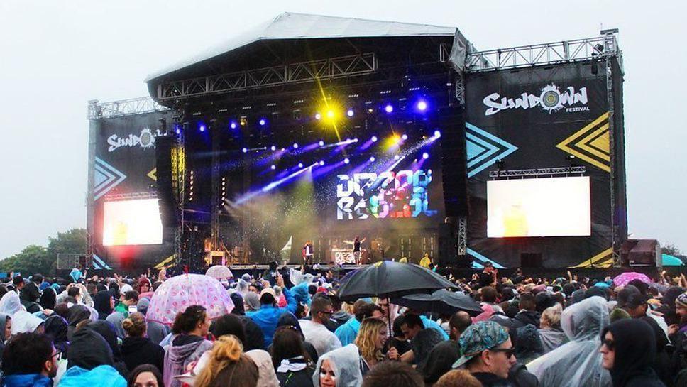 Sundown festivalgoers in rain in front of a stage, with Dizzee Rascal performing. Some are holding umbrellas.