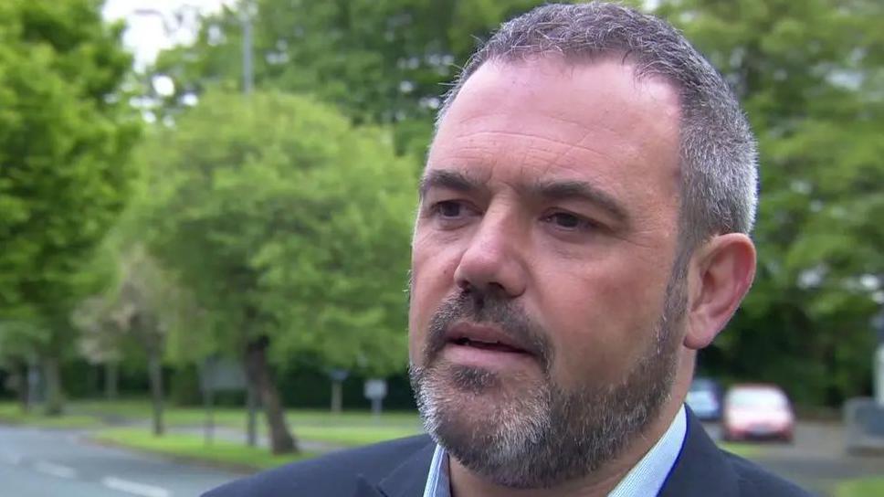 Close up of Paul Foster, who has short hair and a beard. He is standing outside in front of some trees