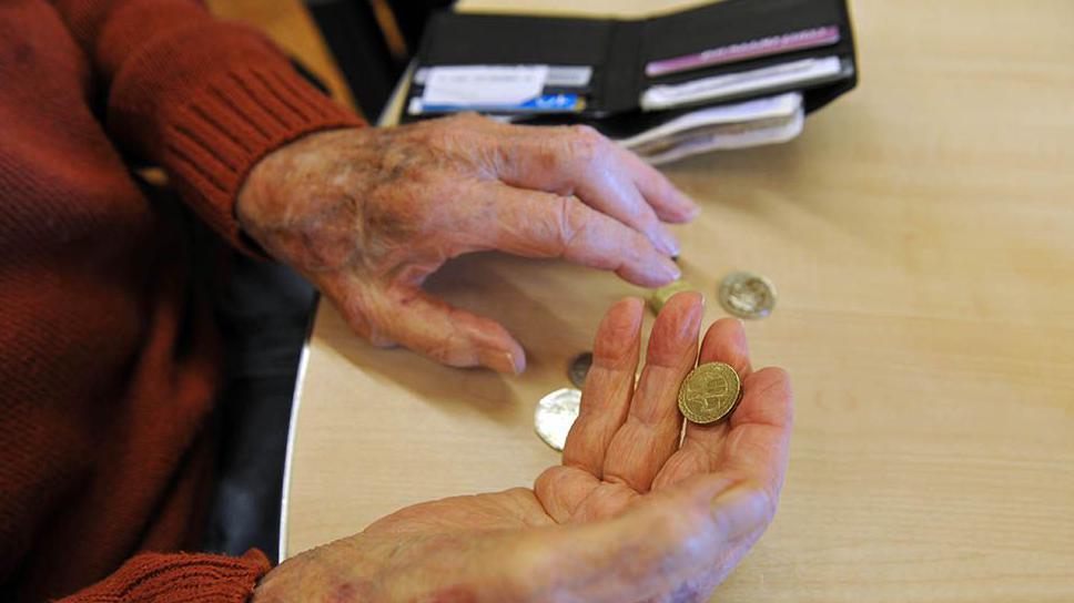 The hands of an elderly person holding a pound coin at a table that has an open wallet in the background