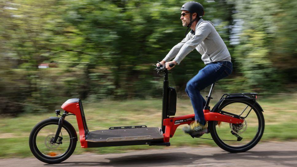 A man wearing a cycle helmet rides an electric cargo bike. He is cycling past trees.