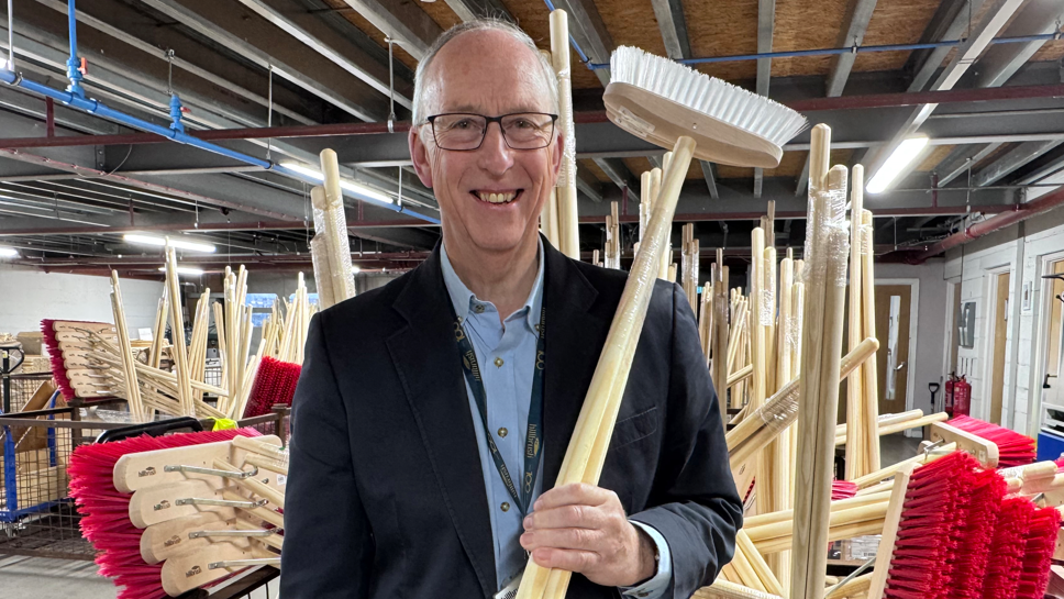 A bespectacled smiling man stands in a large room surrounded by brushes and is holding a brush upside down with his left hand