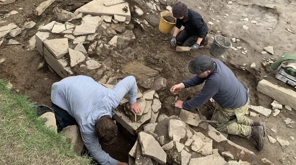 Three people on their hands and knees carefully digging and uncovering stones on the site