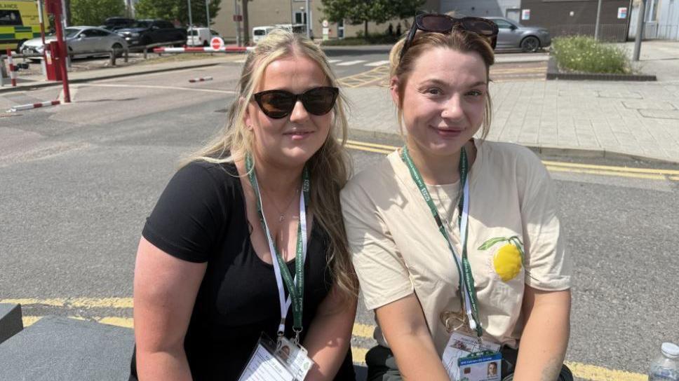 Two women sat on a stone bench in front of a car park. They are both wearing a green and white lanyard. One woman has dark sunglasses on and blonde hair half tied up and the other has dark sunglasses on her forehead with her hair tied back. One is wearing a black top and the other a white top with a lemon on it
