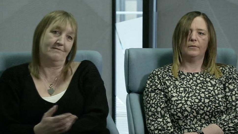 Patricia Gerrard, who has blonde hair and a fringe and is wearing a black top and Samantha Barnes, wearing a green and white top with a floral pattern, look into the camera with a serious expression.