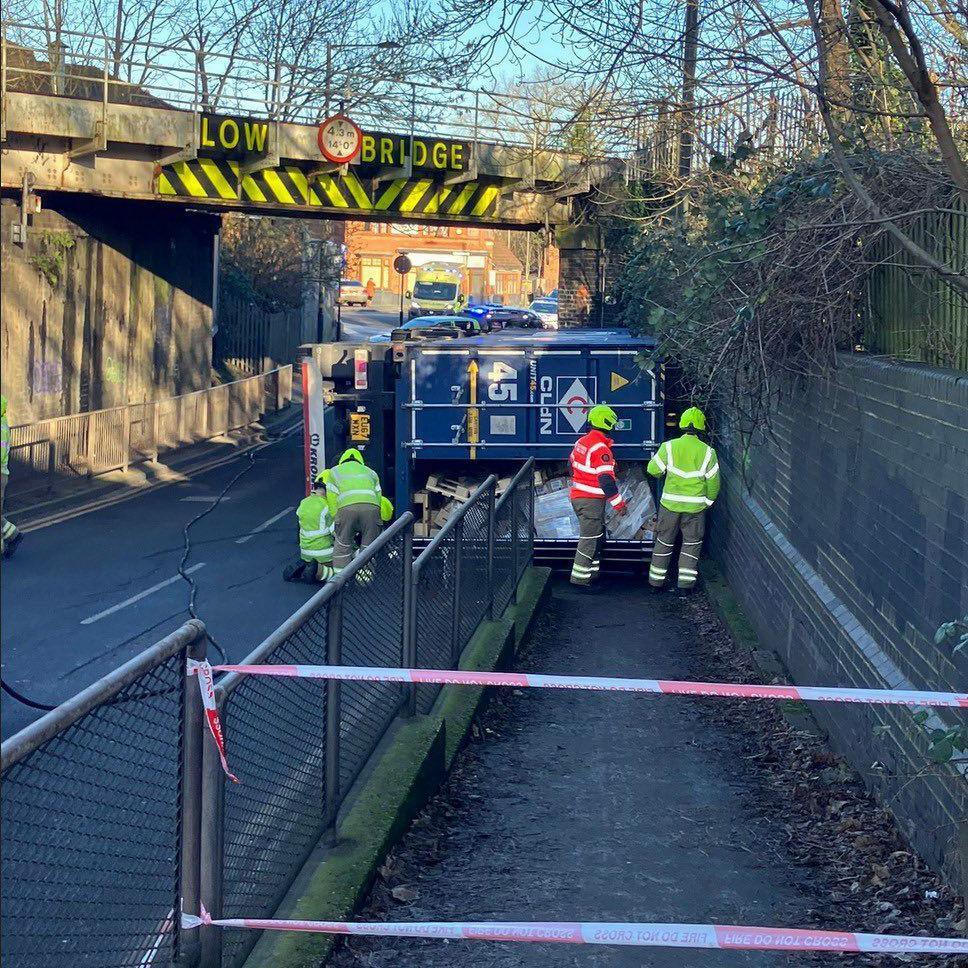 A lorry lies on its side on a road underneath a bridge labelled "LOW BRIDGE". People wearing hi-vis clothing and helmets are inspecting the vehicle and the path on which they are standing has been cordoned off.