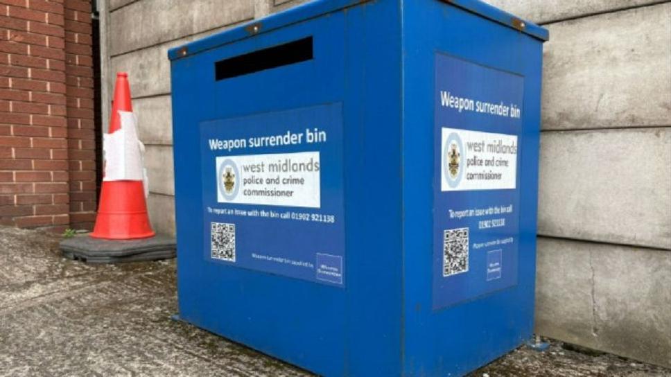 A large blue bin with the words "weapon surrender bin" in white and bar code. A traffic cone is placed next to it and it is located outside in a car park. 