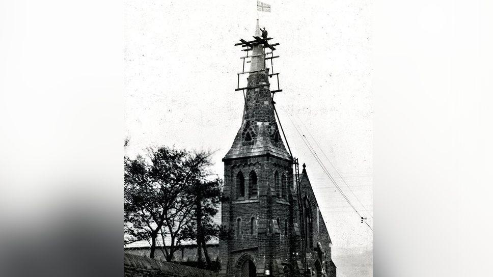 A man stands near the top of a church spire with scaffolding around it after putting a Union Jack flag at the top of it in about 1927.