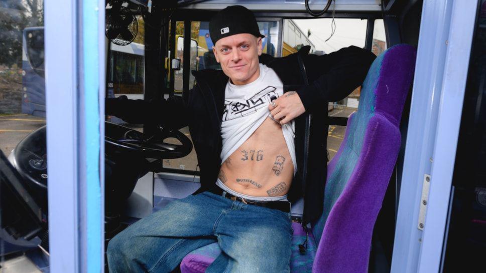 Charlie Chamberlain is sitting in a bus driver seat and lifting his shirt to reveal the number 376 tattooed on his stomach.