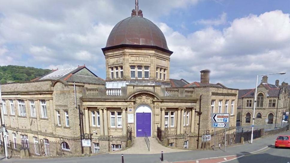 Street view of Darwen Library, a large stone building on a corner plot with a large dome over the main entrance with a purple door, and extensions to each side