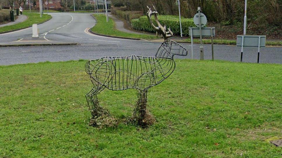 The wire-framed stag sculpture in the middle of a grass-covered roundabout with a road snaking of in the background