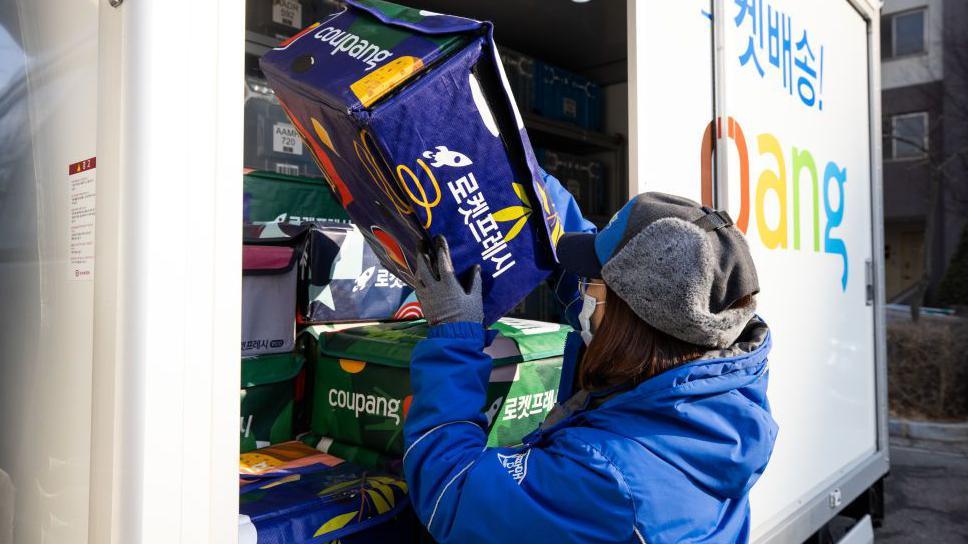 A Coupang employee wearing a protective mask unloads an eco-bag carrying fresh food from a delivery truck in Bucheon