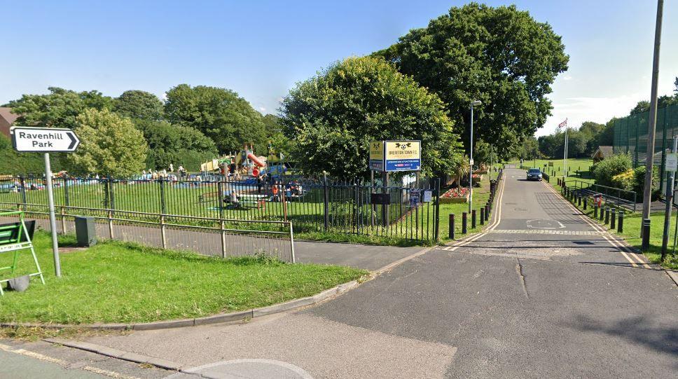 The entrance to a park. A signpost saying Ravenhill Park can be seen on the left, and a long driveway with a single car approaching. A children's playpark is on teh left and a sign for Brereton Town FC. There is a further grass area on the right.