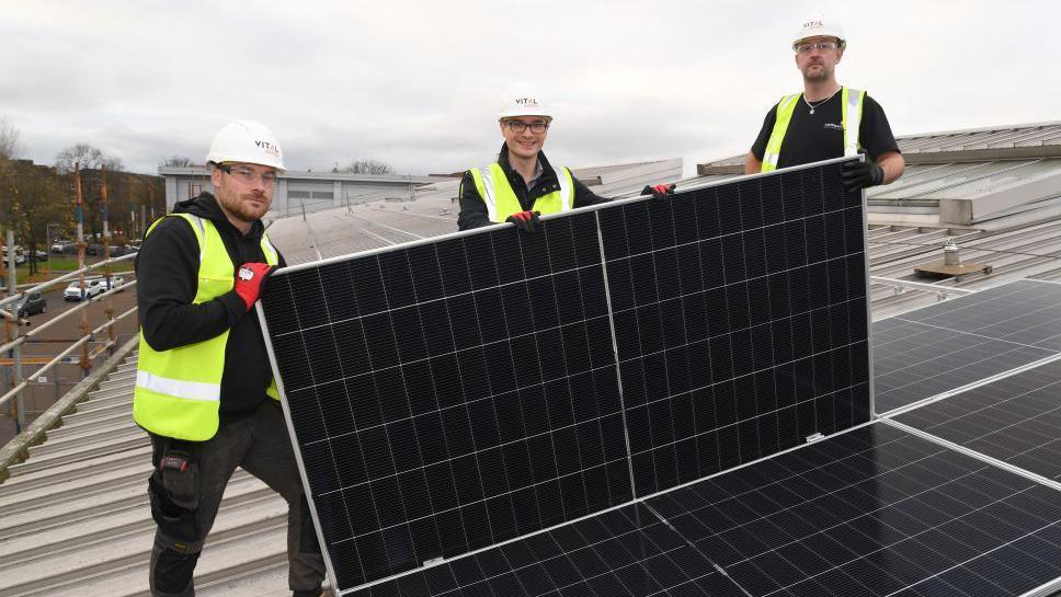 Three men wearing white hard hats and yellow hi-vis jackets holding a solar panel on the roof a school. 