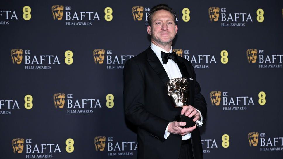 Lol Crawley in black tie attire, holding a BAFTA trophy. He is smiling at the camera and the logo of the EE BAFTA Film Awards adorns the backdrop behind him.