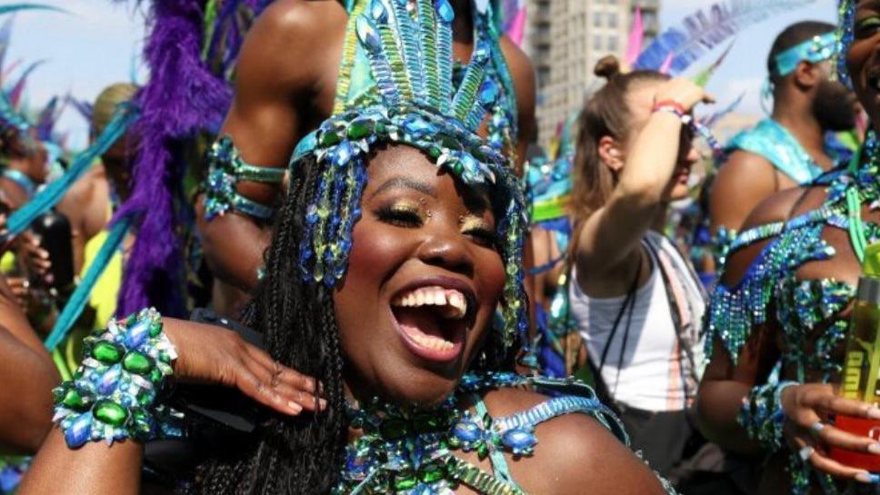 A woman wearing a blue and green sequinned head dress as well as a beaded costume smiles while other people stand in the background at Carnival