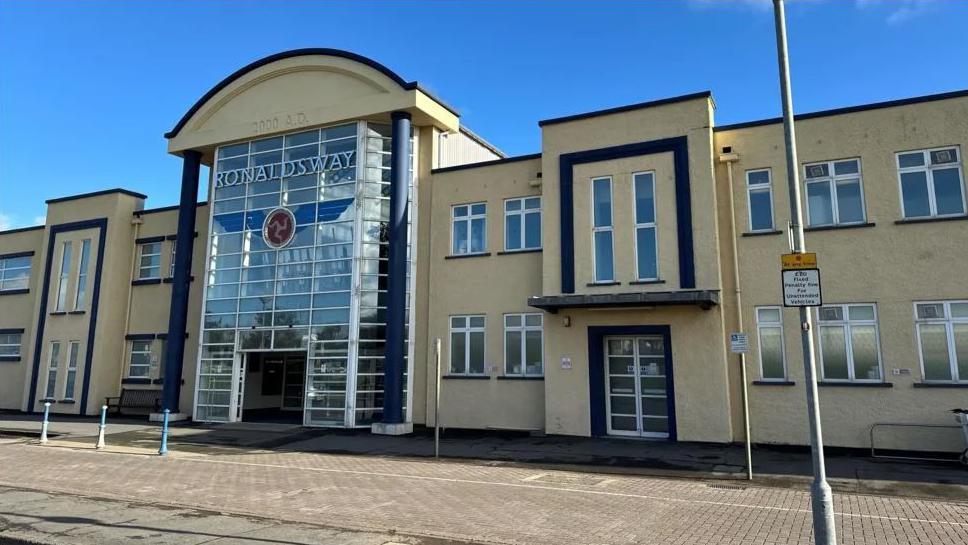 The exterior of Ronaldsway Airport, which is a cream coloured building with lots of windows. There is also tall glass entrance and a sign that says Ronaldsway above the door.