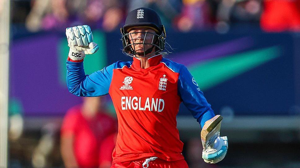 England batter Danni Wyatt-Hodge punches the air in celebration after scoring a century against Sri Lanka. She is wearing red kit, batting gloves and navy blue England helmet.