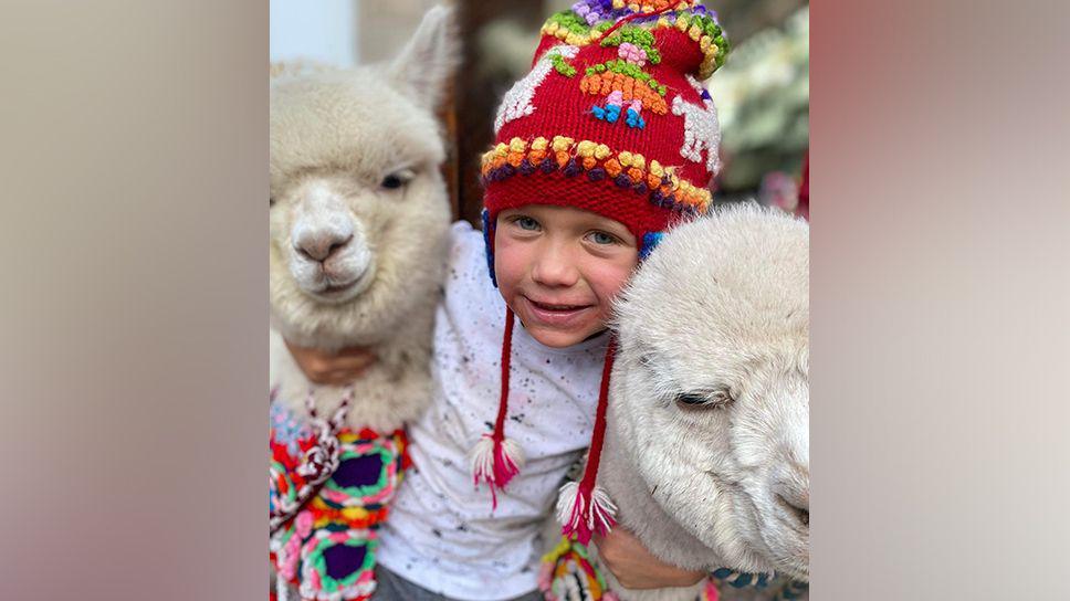 Jamie wearing a red woolly hat and white t-shirt smiling as he hugs two llamas in Peru