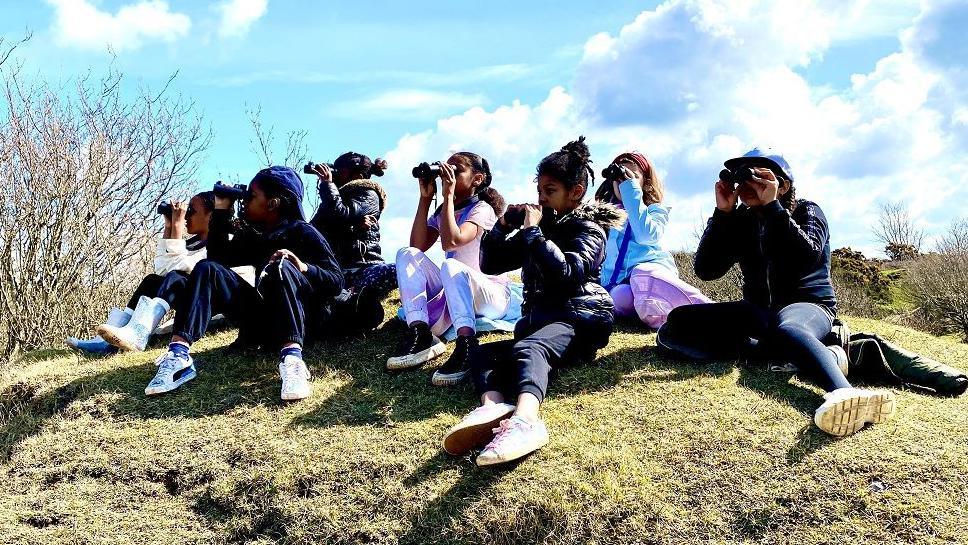 A group of children birdwatching on top of a hill. They are all sitting down on the hill with their binoculars.