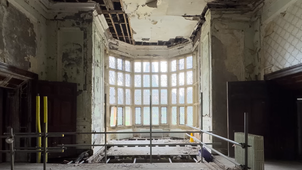 Inside one of the derelict rooms in Hopwood Hall. The image shows a large bay window, scaffolding, and walls stripped of paint and wallpaper. 