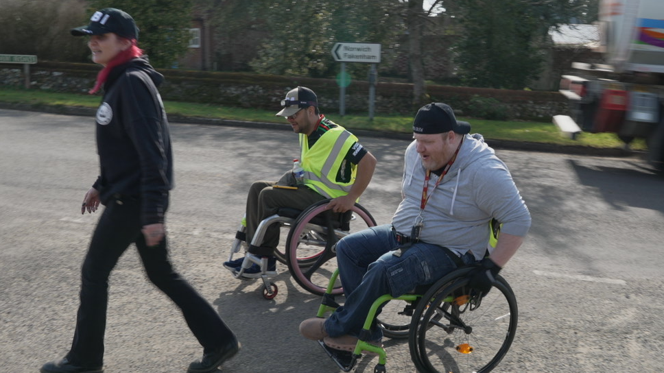 A woman in black clothing and a baseball cap walking across a road, with two men in wheelchairs following behind her, one in hi-vis and dark clothing, the other in a grey hoodie and dark trousers. All three are wearing baseball caps. The blurred image of the back of a moving lorry can be seen to the right, and a road sign for Norwich and Fakenham. 