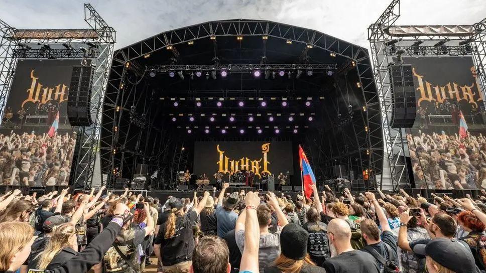 Thousands of fans stand with their arms in the air in front of a large stage at Bloodstock Festival 