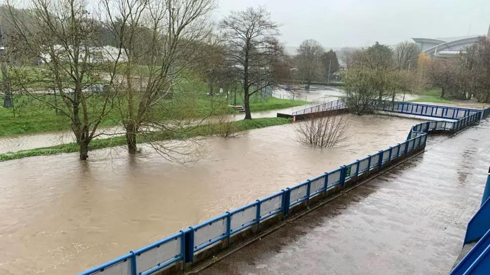 The river running through the NSC grounds at a high level. There are trees and a grassed area on the left and a walkway with blue railings on the right.