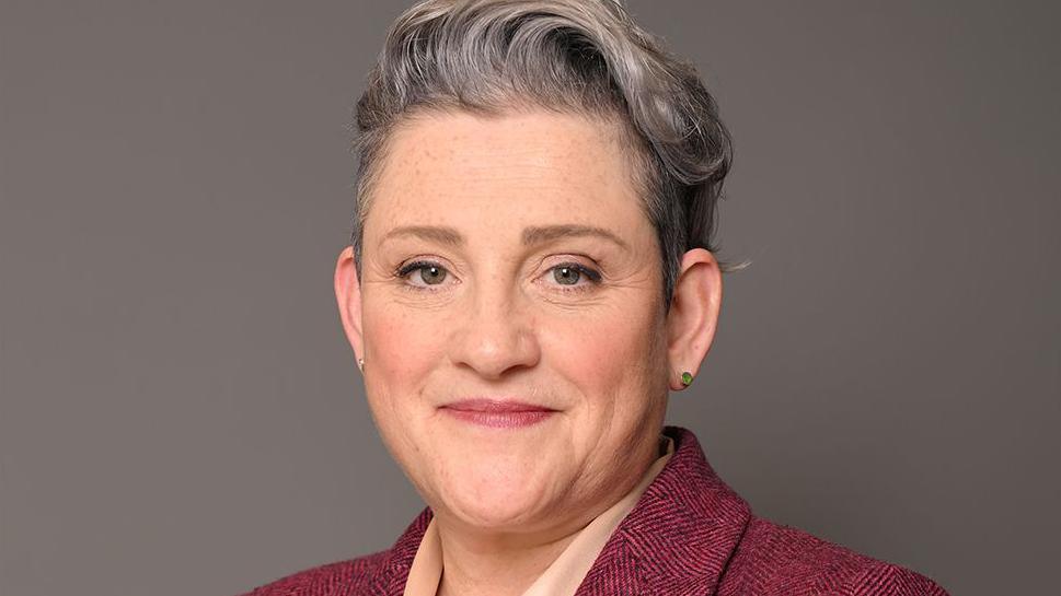 Head shot of Ashley Dalton in front of a plain, dark grey background. The MP is smiling and has short grey hair which is longer on top. She is wearing a burgundy suit jacket and a cream blouse.