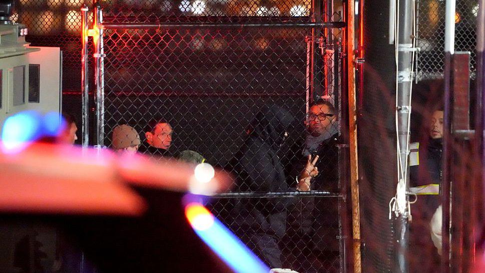 Several people in the distance, including a man dressed all in black, including a black hood, and you can see his hands in the peace sign. A police car is in the foreground.