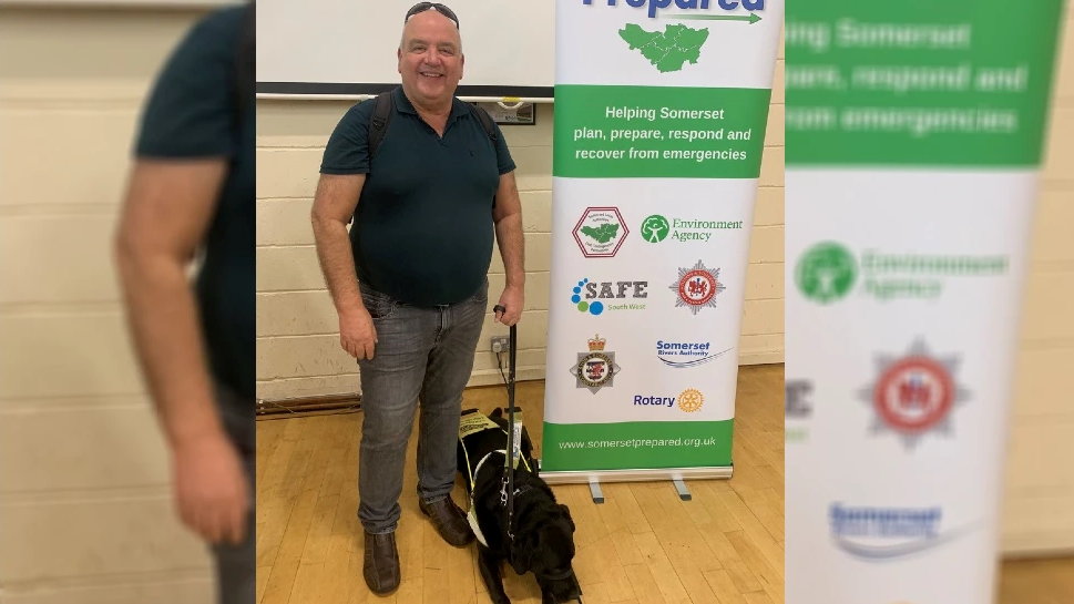 John Hardy in a polo shirt and jeans, smiling, holding his black guide dog who is lying on the floor. He is next to a pull-up banner with Somerset emergency services on it.