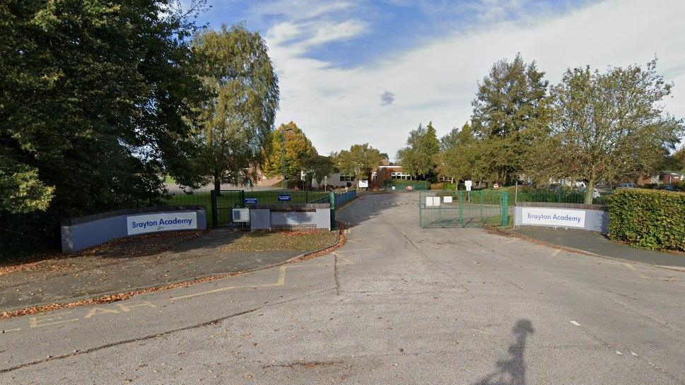 Picture shows the main entrance to Brayton Academy with one green gate closed in the middle of the picture with walls and hedges and trees to the left and right of the entrance gate