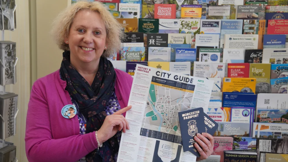 Ruth Smith, Worcester Tourist Information Manager, holds two Worcester Passports and a city guide. She is wearing a pink cardigan, a black patterned scarf and a "History Hound of Worcester" badge.
