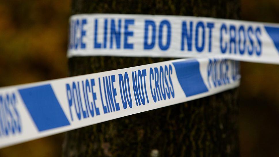 Generic image of police tape wrapped around a tree, it says 'police line do not cross' in blue writing on a white background