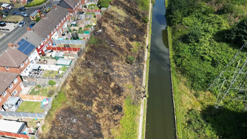 A grassy embankment next to a long strip of canal is seen burnt, turned dark brown and light yellow, with the burnt grass touching residential houses' back garden fences. The other side of the canal is bright green with lots of trees and vegetation, as well as a pylon