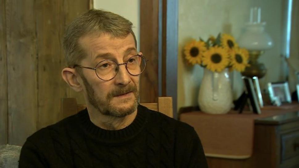 A close-up of a man in a dark jumper, who is wearing glasses and sat in a chair. He is in his living room and behind him is a brown unit with a vase of sunflowers on the top.