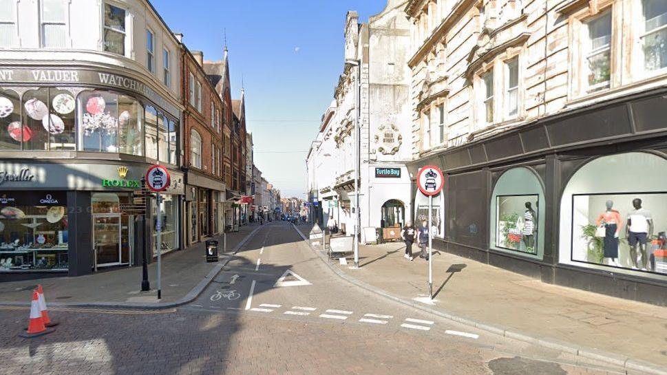 Gold Street, showing no access for cars or cycles. A jeweller's shop is visible on the street corner to the left, and a dress shop to the right. Two road cones can be seen to the left.  Other shops are visible in the distance.