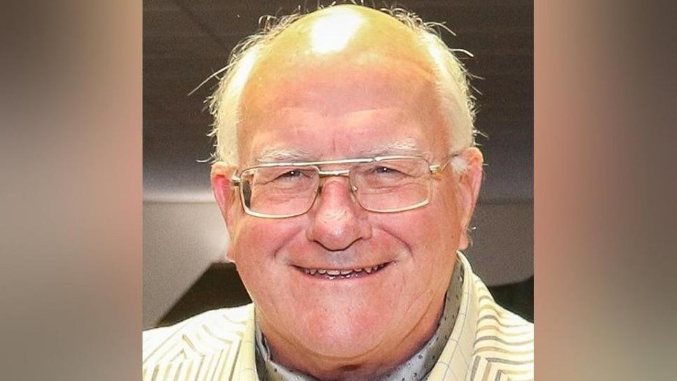 Kenneth Wright, smiling and wearing glasses. He has a yellow jacket that includes grey and brown stripes.
