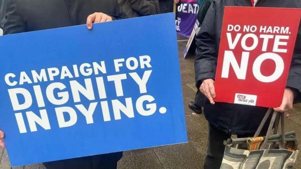 A person holding a blue sign with the word Campaign for Dignity in Dying, alongside another holding a red sign says Do No Harm. Vote No.