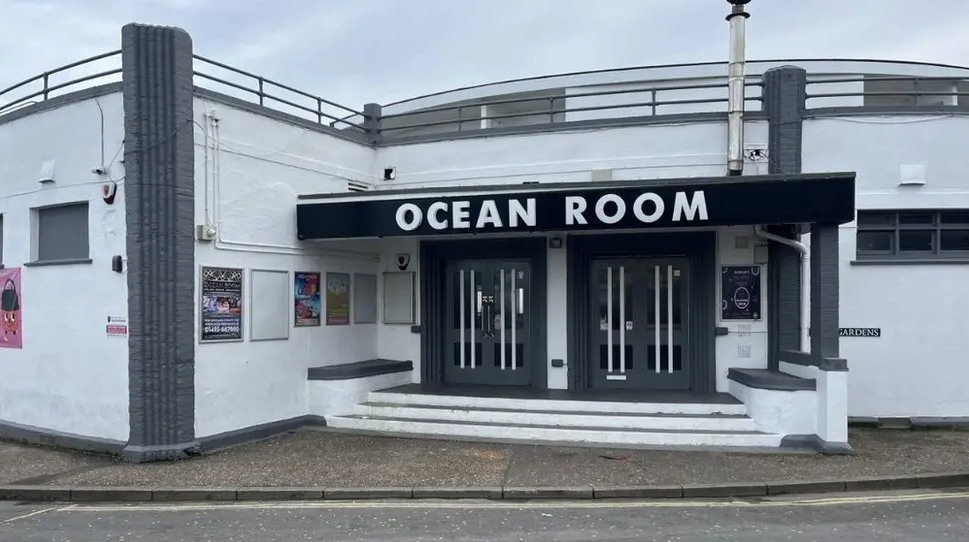 The Ocean Room, a white rotunda building, with grey columns, built in the Art Deco style. The building features entrance doors with signage reading "Ocean Room" above, and to the left is a castellated staircase that leads to the roof terrace. 