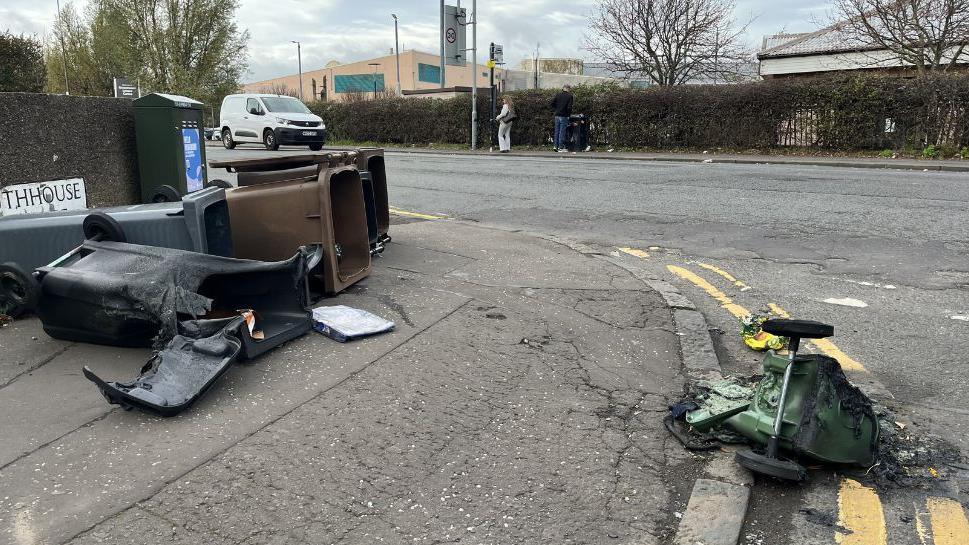 Melted bins on the side of Captain's Road. A green bin is next to the kerb on double yellow lines and has been badly melted. Its two wheels are attached, with one sticking in the air. A black bin is partially melted on the pavement. It is next to another black bin and two brown bins which are unaffected. 