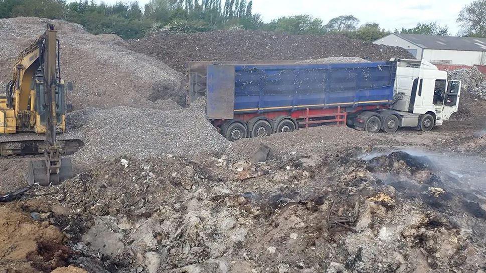 A lorry and digger at the site surrounded by piles of burnt waste.