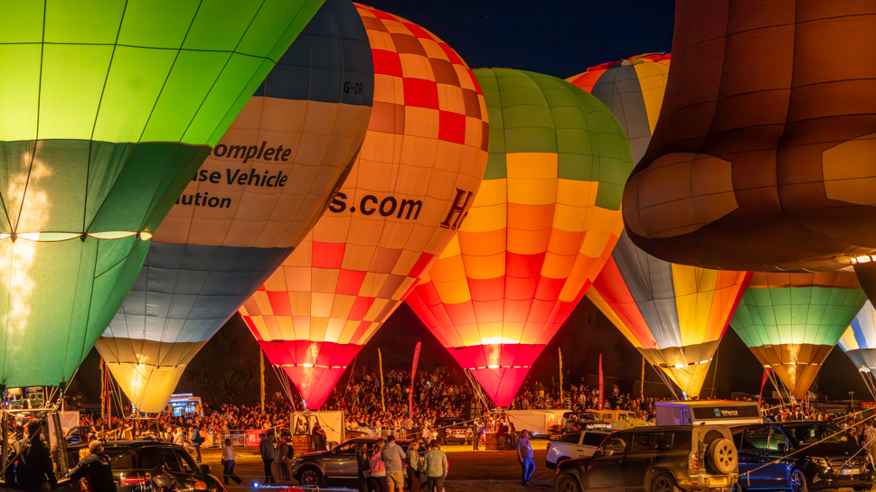 About eight colourful hot air balloons are inflated next to each other and tethered to the ground. It is dark and the burners are lighting up the balloons. A crowd is gathered in the background.