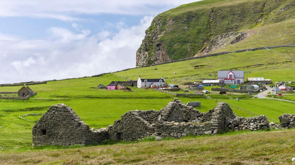 Remote farmhouse on the Shetland island of Foula, with ruins of a farm building in the foreground, amid grassy fields, and rocky high cliffs in the background.