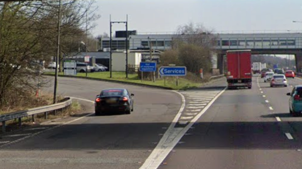 The slip road to a motorway services, with three lanes of traffic to the right of the services, and a covered bridge over the road