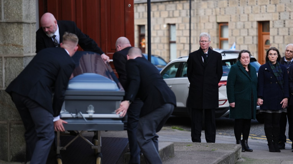 Four men carry a grey coffin up a few steps through a doorway on the left of the photo. On the right klenny Mackaskill and two women are standing. All are dressed formally in black.